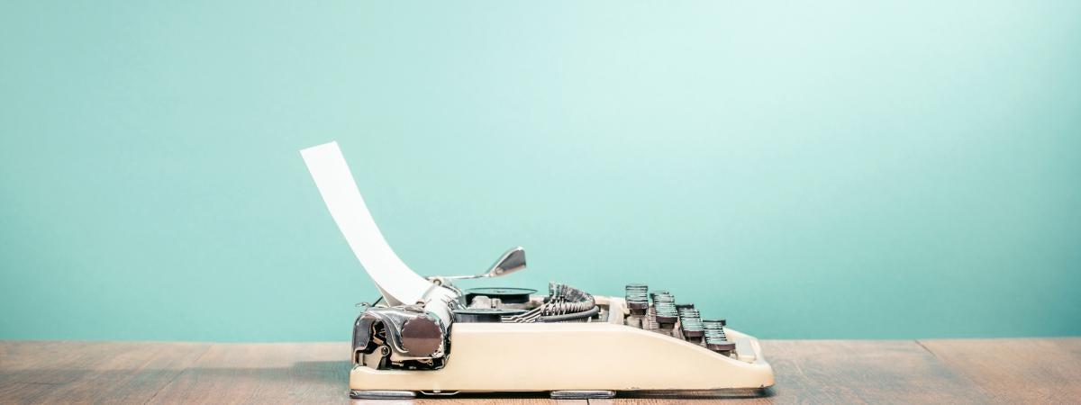 A small white typewriter sits on a table against a teal background